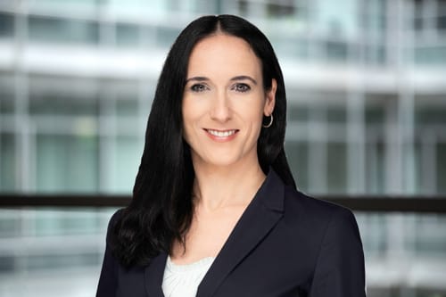 Melanie Kramp-Gerstner im Portrait-Format vor einem verschwommenen Glas-/Stahlhintergrund.